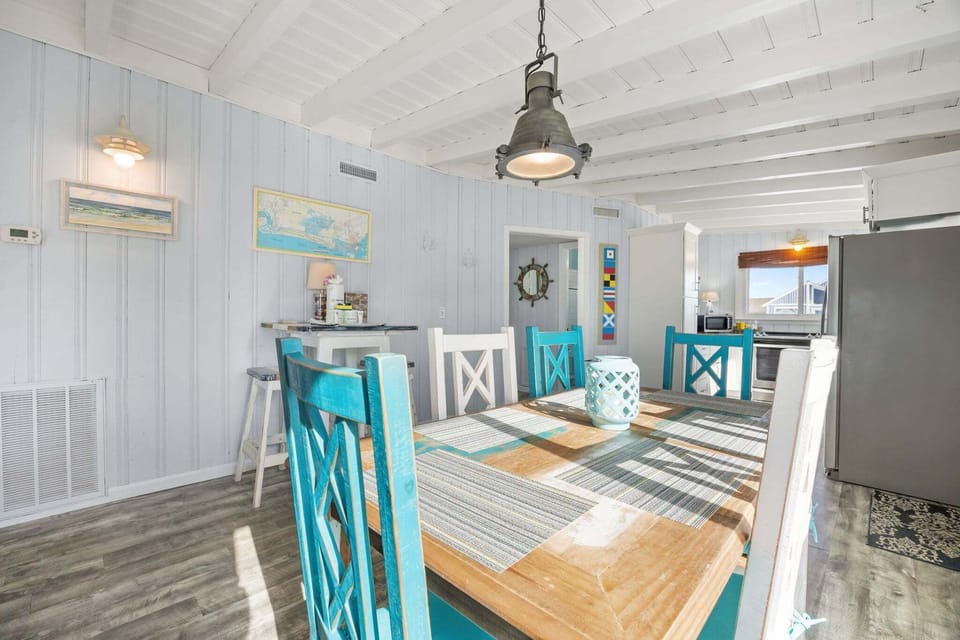 Bright dining room with a wood table, turquoise and white chairs, coastal wall art, beamed ceiling, pendant light, and open view into the kitchen.