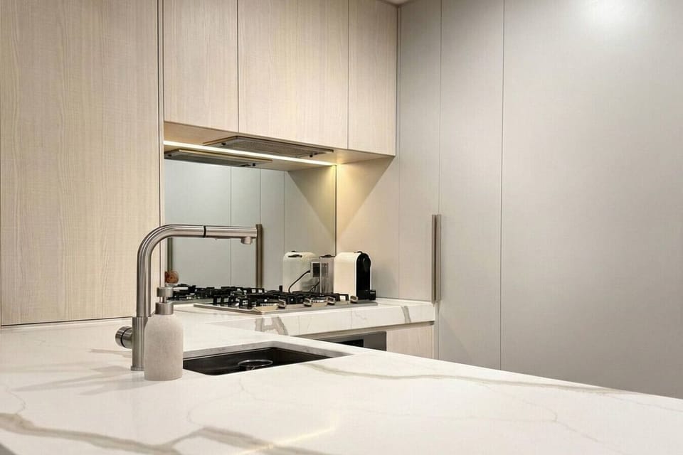 A sleek kitchen with a marble benchtop, brushed steel tapware, gas cooktop, mirrored splashback, and warm timber cabinetry. The Nespresso machine and toaster are set out and ready, making mornings straightforward for guests.