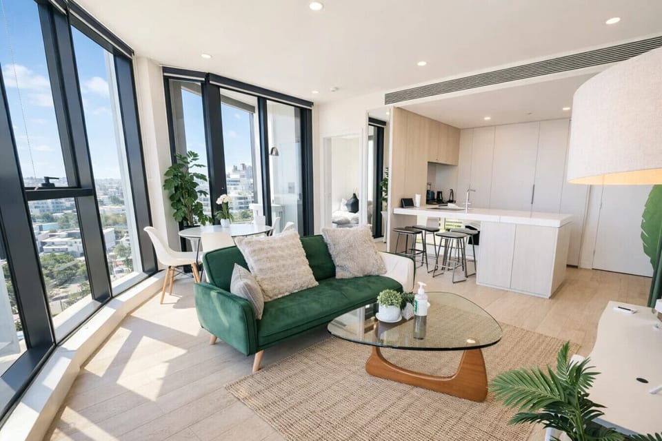 Sun-filled living space with a green velvet sofa, glass coffee table, and indoor plants, flowing into a white kitchen with breakfast bar. Floor-to-ceiling windows flood the room with natural light and suburb views.