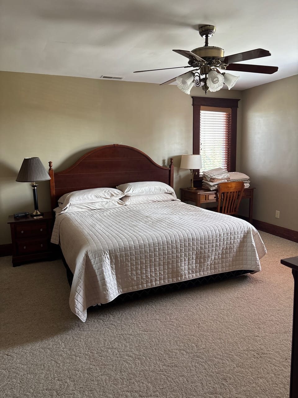 Cozy, carpeted bedroom with king bed, ceiling fan, and sunny workspace