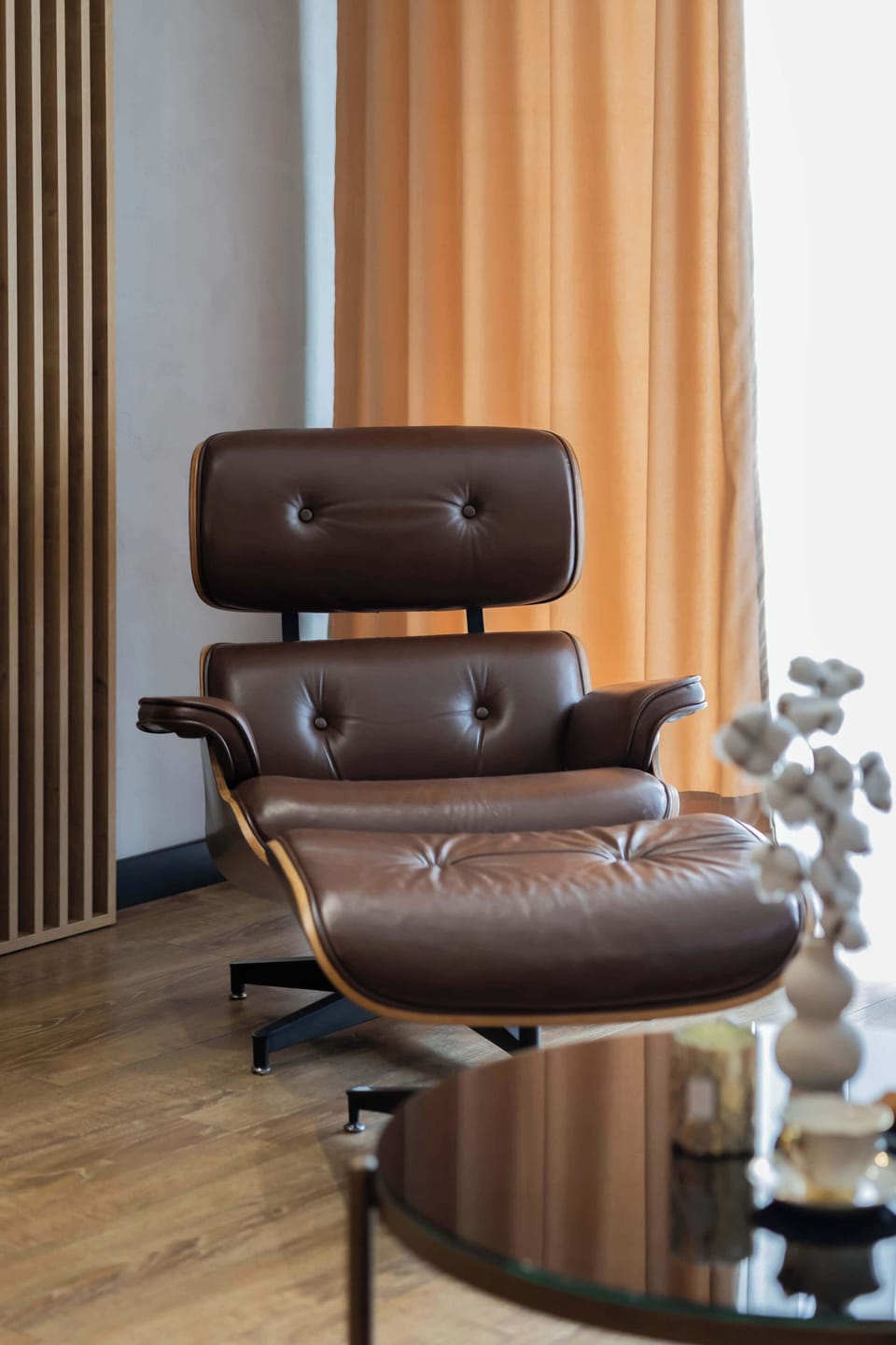 A modern armchair with a matching footrest placed next to a floor lamp, creating a perfect reading corner. Warm curtains and soft lighting make the space feel comfortable and inviting.