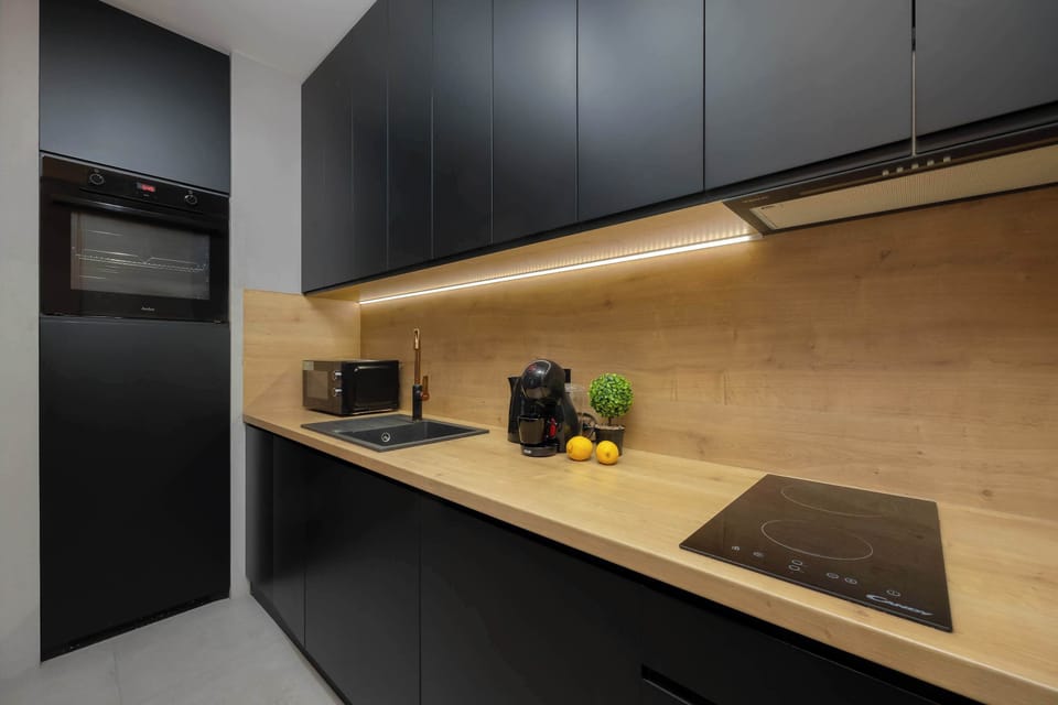 A minimalist kitchenette with clean lines and handleless cabinets in dark tones. Warm under-cabinet lighting highlights the wooden backsplash and worktop, giving the space a cozy feel.
