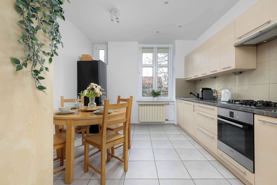 The dining area in the kitchen is illuminated by a window and features a classic wooden table with matching chairs. Green trailing plants on the shelves add a fresh and lively feel to the room.