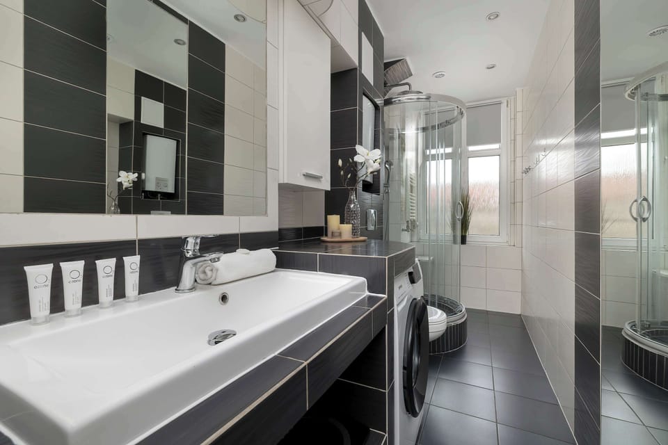 The bathroom displays a modern aesthetic with black and white wall tiles and a sleek white vanity unit. A glass shower cabin and a washing machine are neatly integrated into the space.
