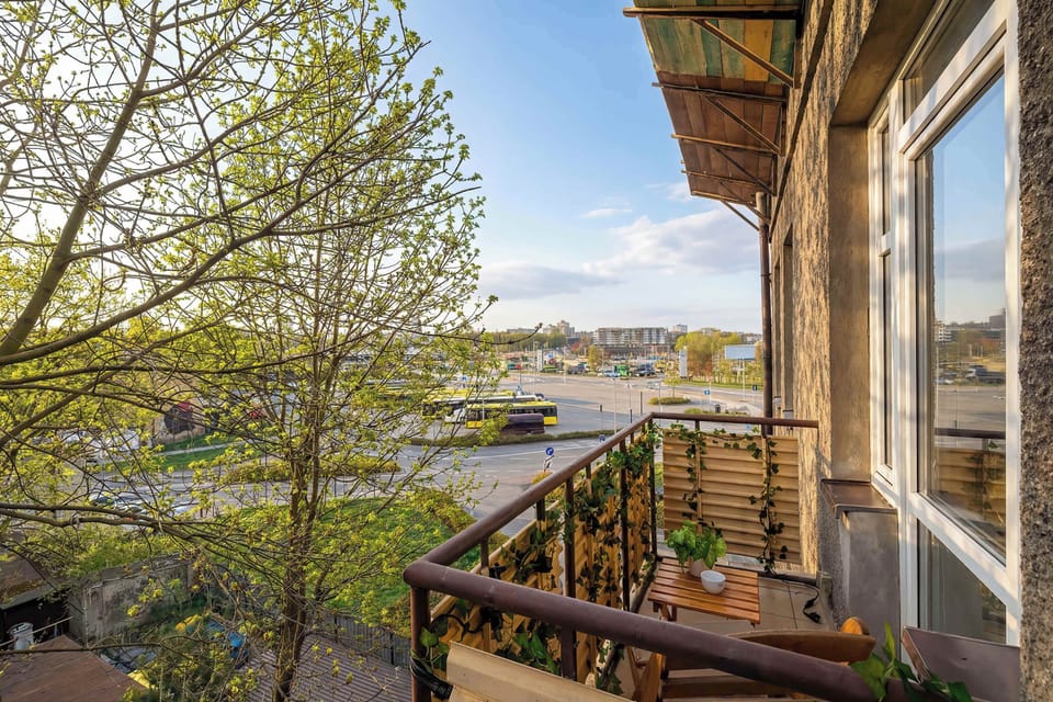 A view from the balcony looks out over the surrounding neighborhood, showing green trees and urban life. The balcony itself is well-maintained with wooden accents and potted plants.