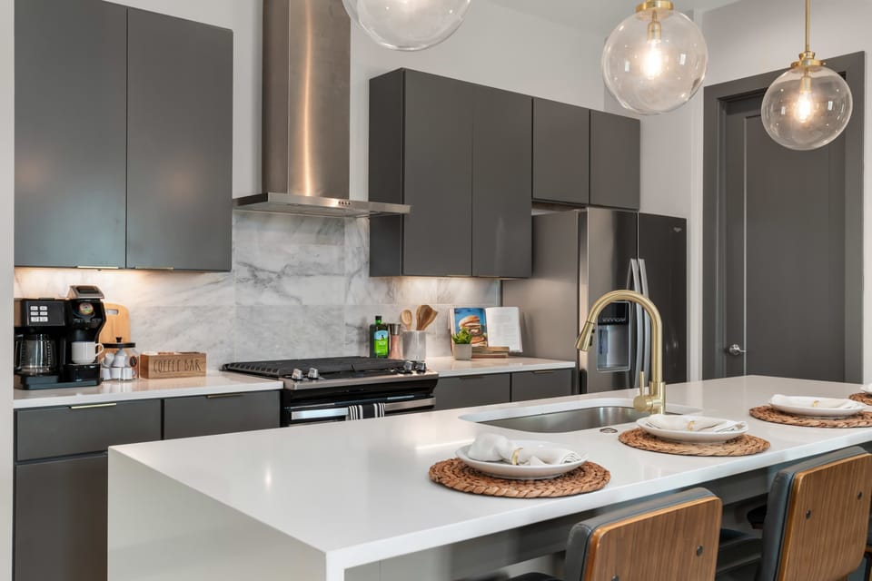 The chef's kitchen features graphite flat-front cabinetry, a marble slab backsplash, a gas range, a stainless steel range hood, a French door refrigerator, a quartz island with bar seating, and brass fixtures throughout.