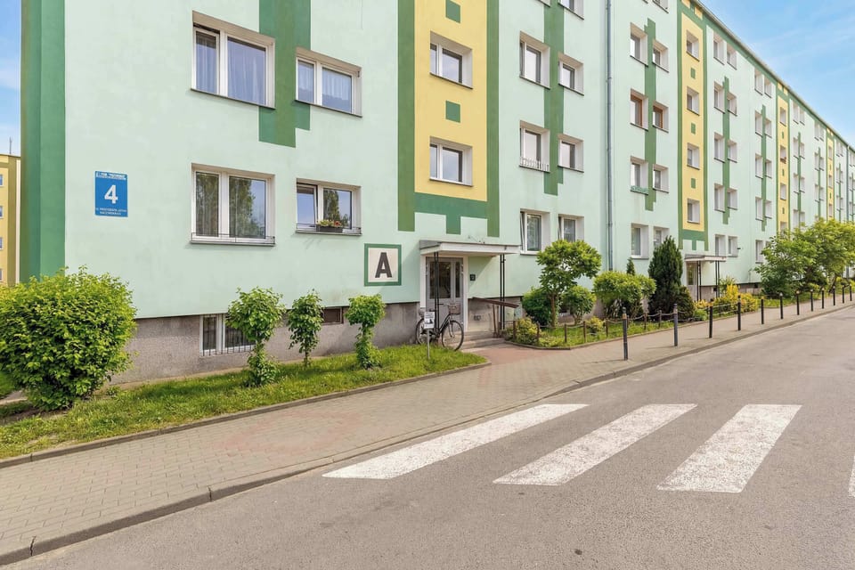 The exterior of the apartment building is painted in light green and yellow tones with numbered entrances. A paved sidewalk and small green bushes run along the front of the structure.