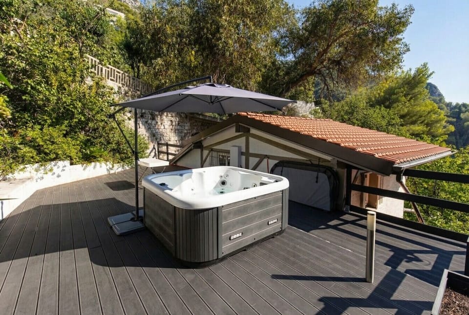 An outdoor hot tub on a dark composite deck, sheltered by a large cantilever parasol, with a terracotta-roofed outbuilding and hillside trees in the background.