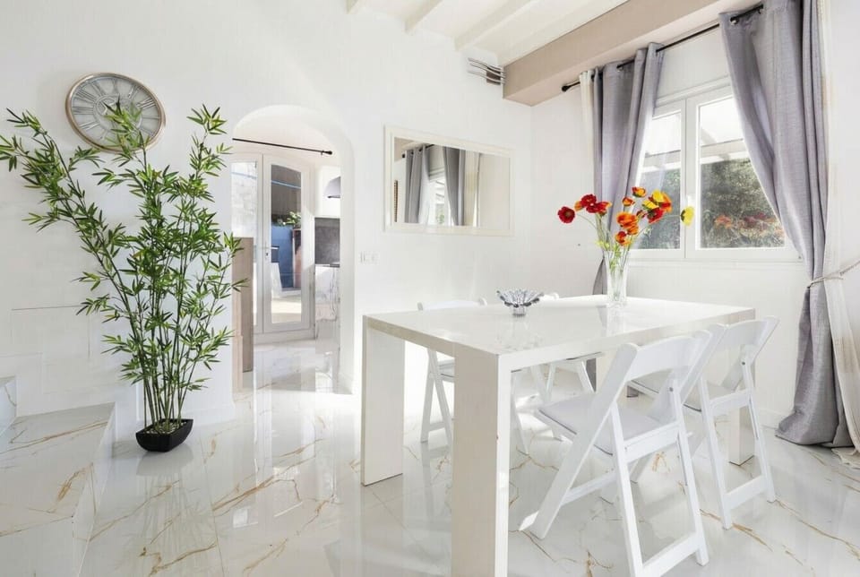 The dining and living room from a different position, showing the white table, bamboo plant, wall clock, grey curtains, and polished white marble floor.