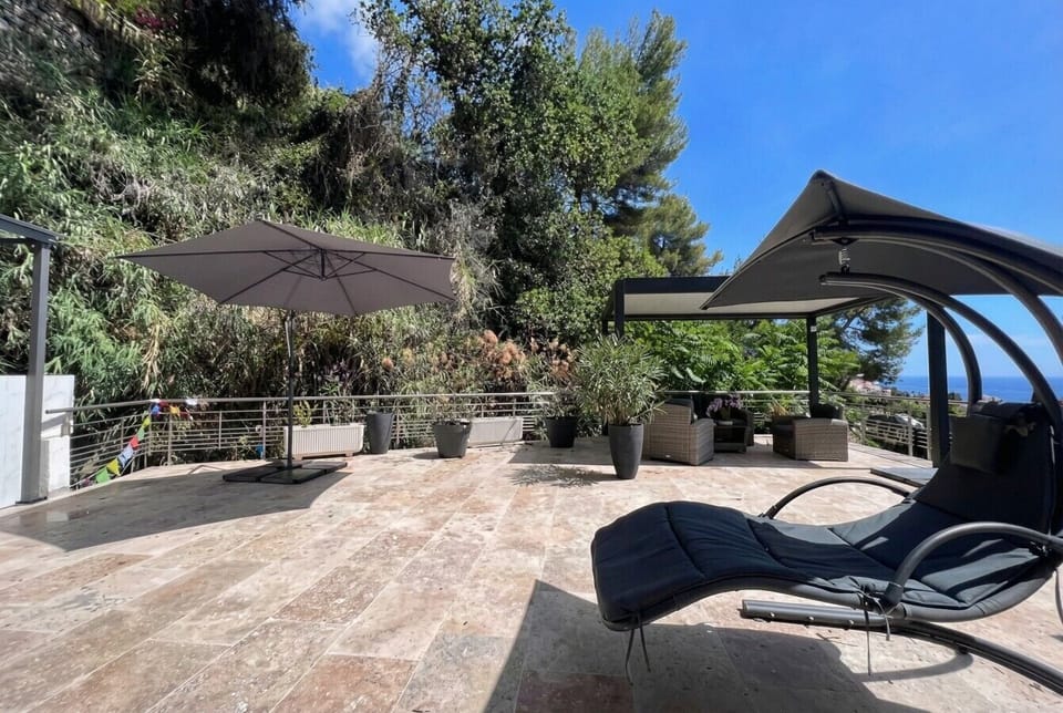 A stone-paved sun terrace with a cantilevered lounge chair, a large parasol, potted plants, and a glimpse of sea at the railing.