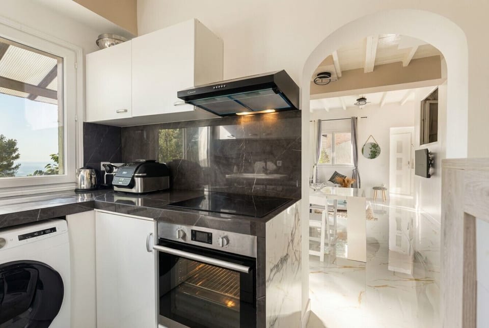 A compact kitchen area with dark countertops, a built-in oven, a washing machine set under a white cabinet, and a rounded archway leading through to a white interior.