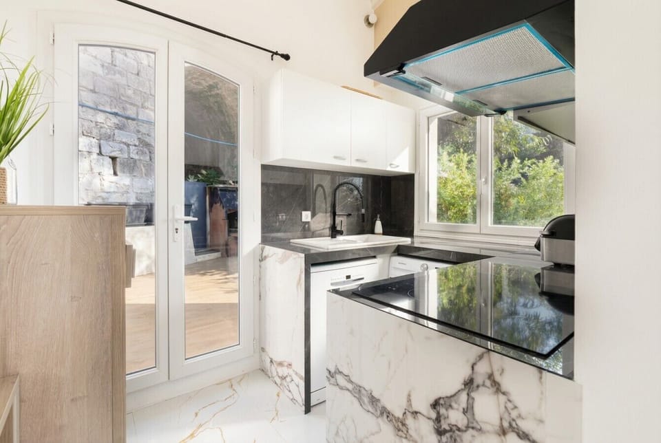 A kitchen counter detail showing a dark surface, marble-veined splashback, and exterior greenery visible through glazed doors behind.