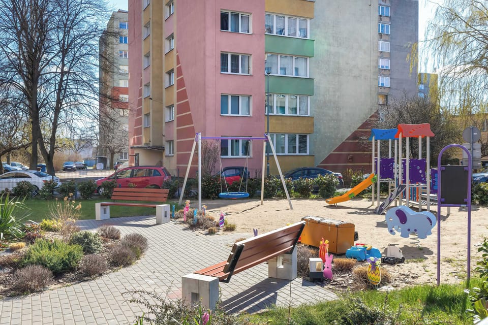 An outdoor playground features a variety of colorful slides, swings, and climbing frames for children. A park bench sits in the foreground, overlooking the play area and the nearby apartment blocks.