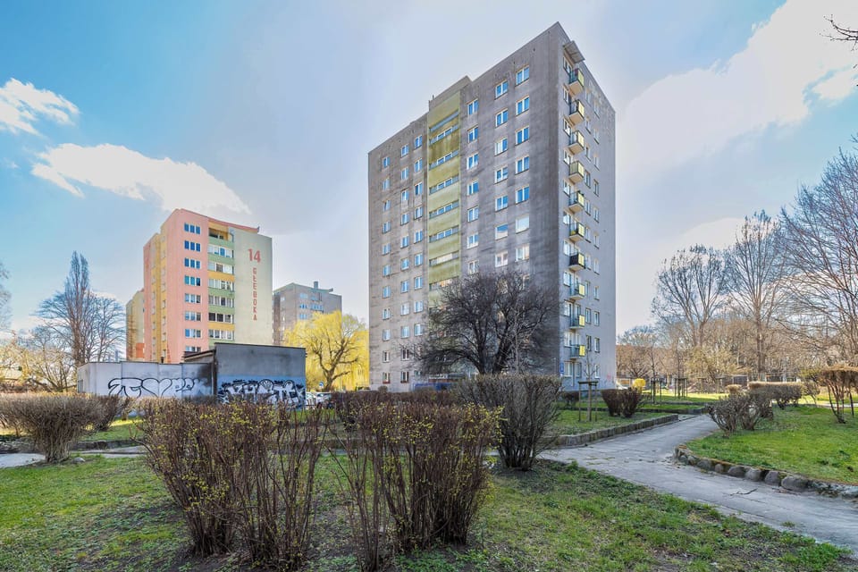 The exterior of the apartment building is shown against a bright blue sky, featuring a mix of green and grey facade panels. Manicured hedges and green lawns surround the base of the high-rise structure.