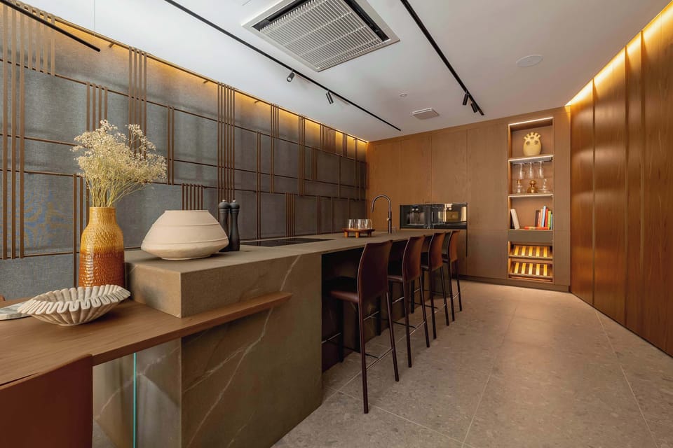 A stylish bar area with a long wooden counter and high bar stools. Warm lighting and wooden finishes create a cozy, upscale atmosphere, while decorative shelving adds character to the space. 