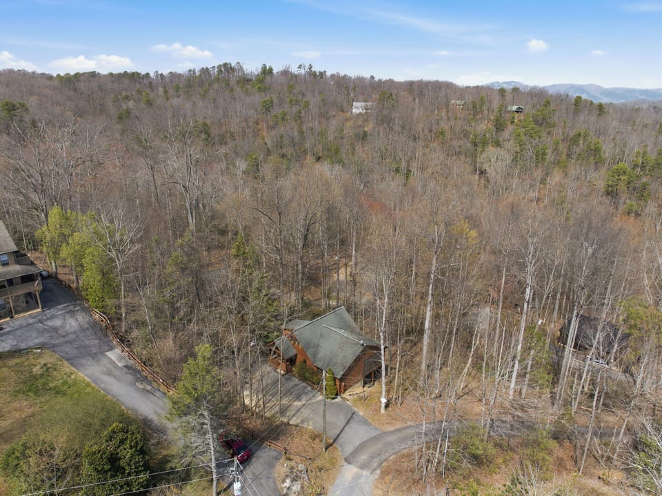 Aerial view of a secluded mountain cabin nestled among rolling forested hills with distant mountain peaks on the horizon.