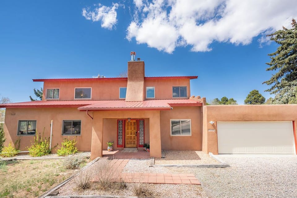 Adobe-style vacation home with distinctive red metal roof and attached garage, set among mature trees under New Mexico's brilliant blue sky.