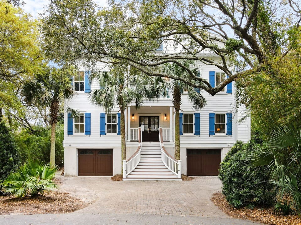 Charming coastal home with blue shutters nestled among palm trees and mature oaks, featuring twin garages and welcoming front entrance.