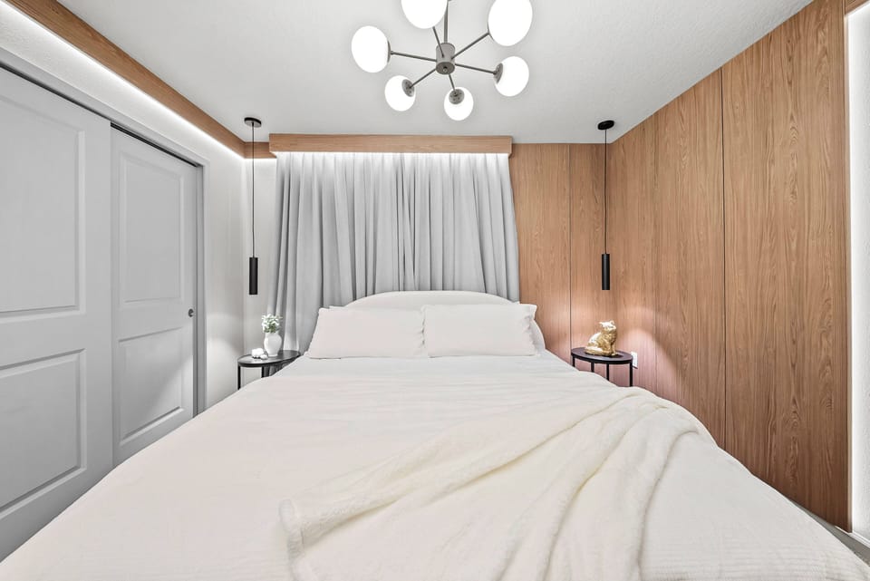 This bright and airy bedroom invites relaxation with its soft white bedding and cozy warm wood paneling. A contemporary light fixture adds a gentle glow, creating a tranquil atmosphere perfect for unwinding.
