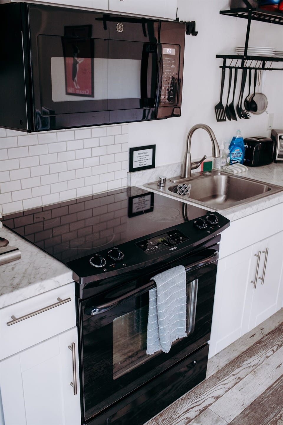 The kitchen has a sleek black electric cooktop set into a marble-inspired countertop with white cabinetry. A stainless steel sink with a high-arc faucet and a white subway tile backsplash complete the modern, functional design.
