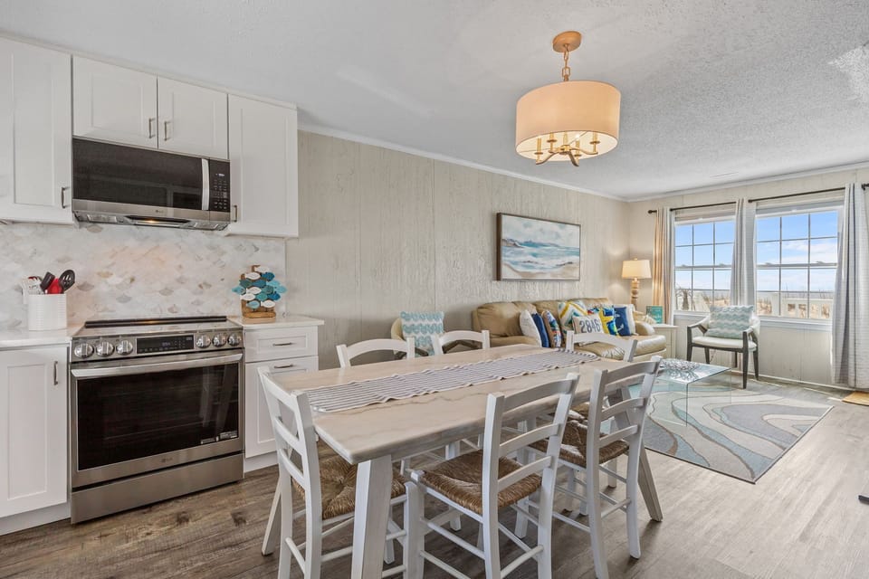 Bright white kitchen with stainless appliances, marble-look table, six chairs, modern lighting, wood-look floors, and stair access nearby.