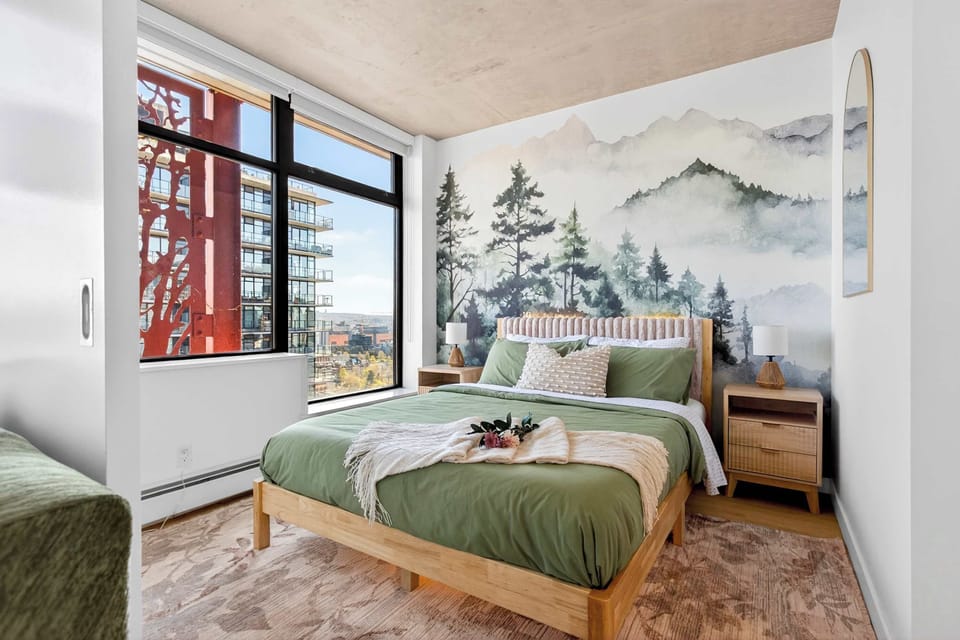Bright and airy bedroom with a custom nature-inspired mural, sage green linens, and large windows offering an urban view of the surrounding architecture.