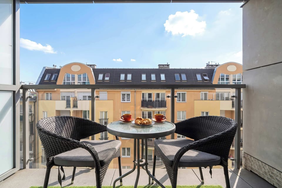 A private balcony is furnished with two black wicker armchairs and a small round table. It offers an elevated view of the surrounding architecture under a clear blue sky.