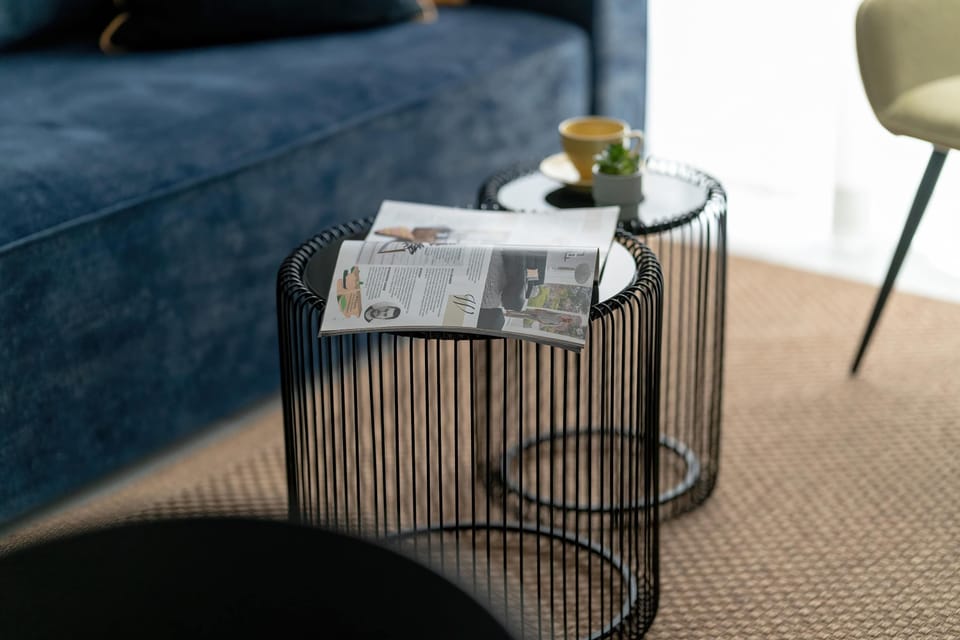 Two nested wire-frame coffee tables sit on a natural jute rug, adding a modern and light aesthetic to the center of the room. Magazines and a small bowl are placed on top, suggesting a space for relaxation.