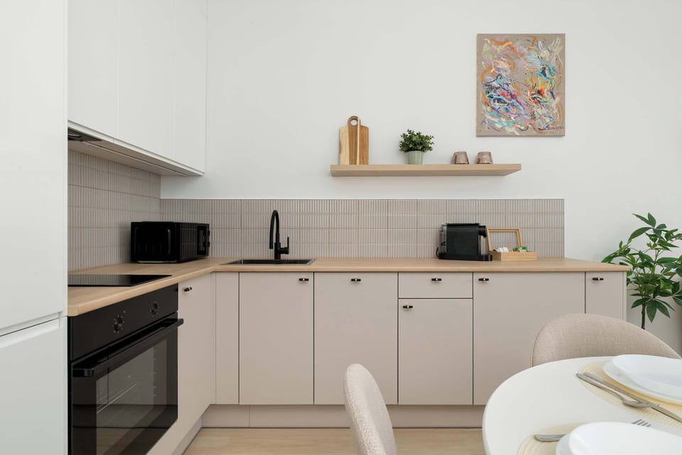 The kitchen wall is adorned with a simple wooden floating shelf holding decorative items and greenery. Underneath, a black sink and faucet provide a sharp contrast to the light wood countertop.