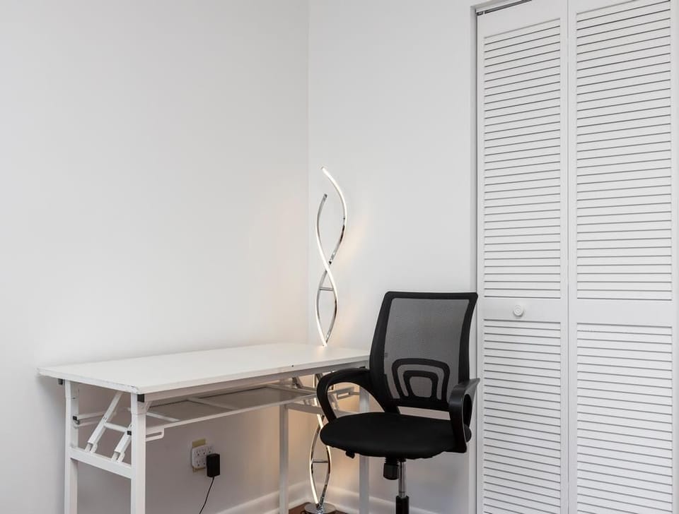 Work space with a white metal desk, black mesh office chair, and sculptural LED floor lamp by louvered closet doors.