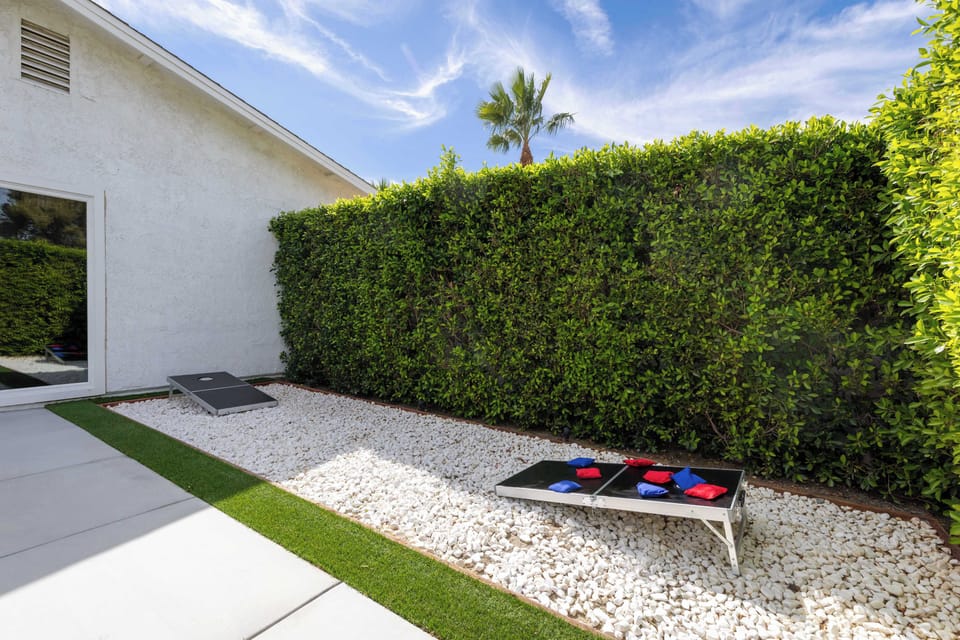 Private cornhole lane tucked along the hedged side yard with white gravel — the group tournament nobody planned but everyone plays.