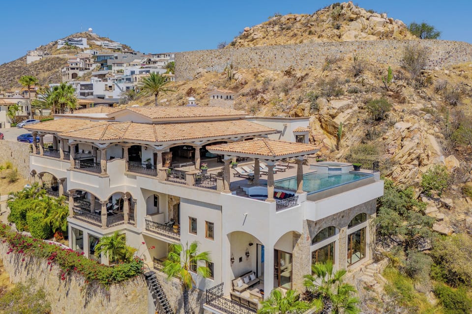 Stunning hillside villa showcasing Mediterranean architecture with rooftop pool amid dramatic desert landscape and neighboring coastal community.