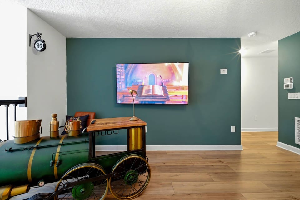 Experience a stylish living area with a rich green accent wall and warm wooden floors that create a cozy vibe. A unique vintage train decor piece adds charm, while the wall-mounted TV ensures your entertainment needs are met.