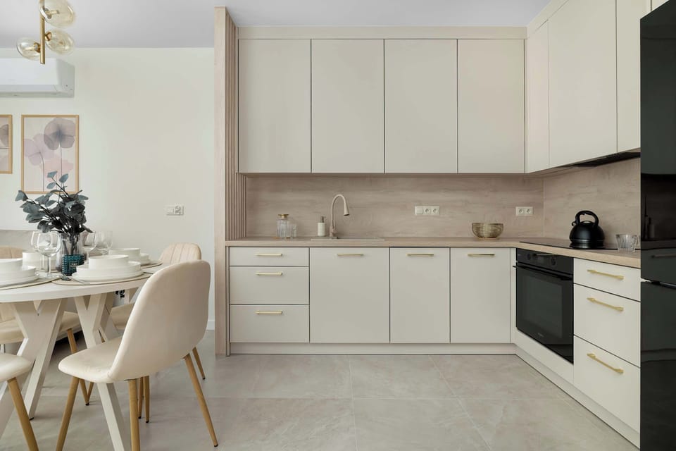 A compact apartment living area features a corner kitchen with cream cabinetry, dark countertops, and a black refrigerator. The space also includes a dining set with four chairs and wall art.