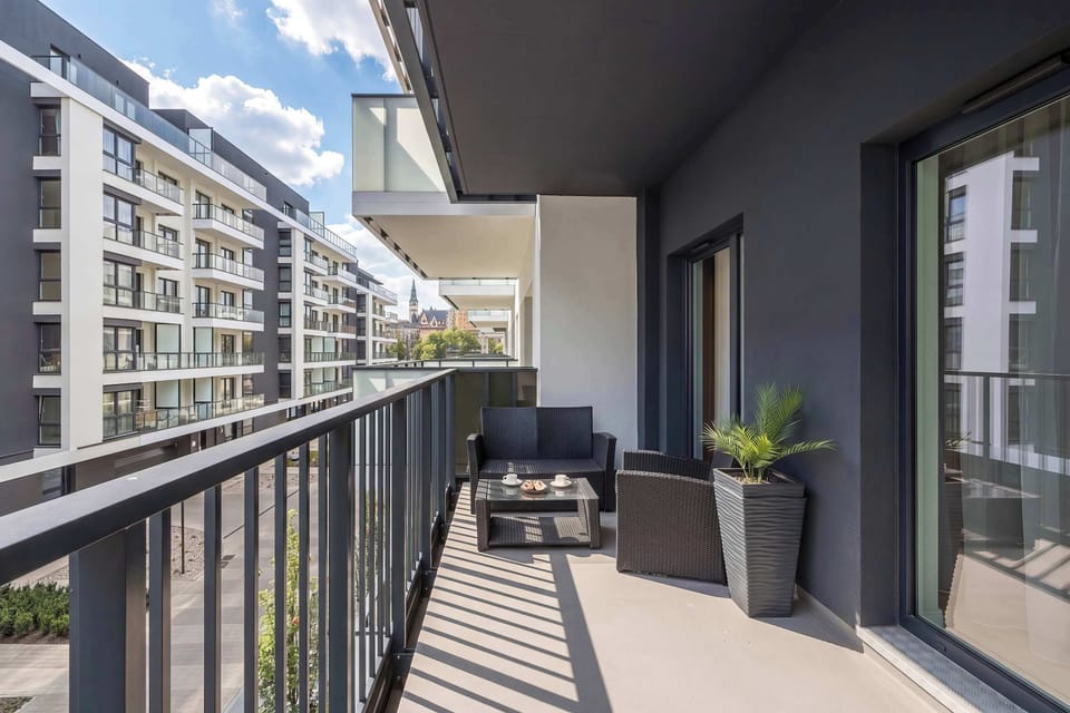 A long balcony with outdoor seating and a view of neighboring buildings. Potted plants add a touch of greenery.