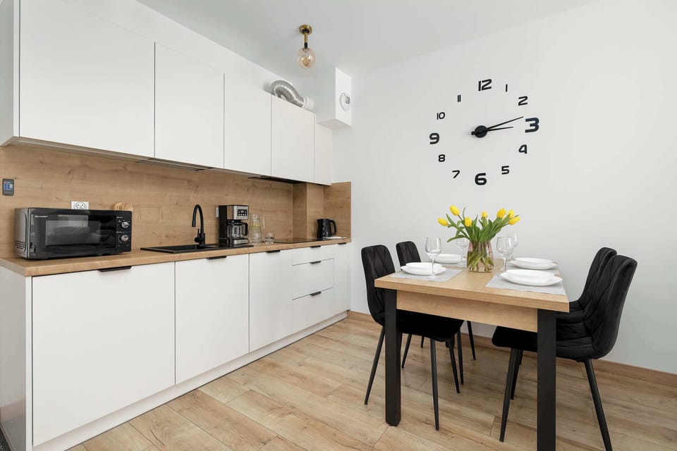 This kitchenette area shows white minimalist cabinetry paired with a light wood countertop and a small dining table with black chairs. A large, decorative clock face is applied directly to the wall, serving as a unique focal point.