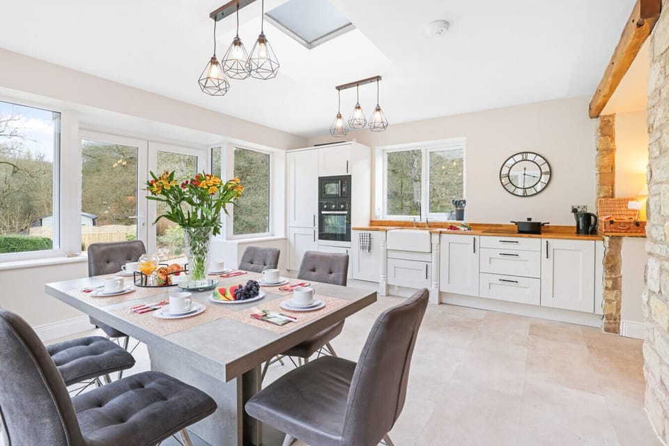 This bright, contemporary kitchen and dining area features white cabinetry, a skylight, and a large dining table set for breakfast, all illuminated by stylish geometric pendant lights.