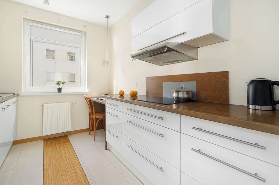 The kitchen perspective highlights a long wooden countertop and a modern induction cooktop. White upper cabinets and a large window ensure the workspace feels open and airy.