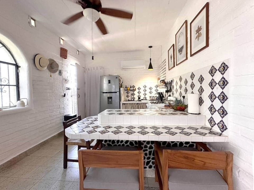 Open kitchen and dining area with a two-tier tiled counter — a bright, connected space with AC, ceiling fan, and a full-size fridge for everyday comfort.