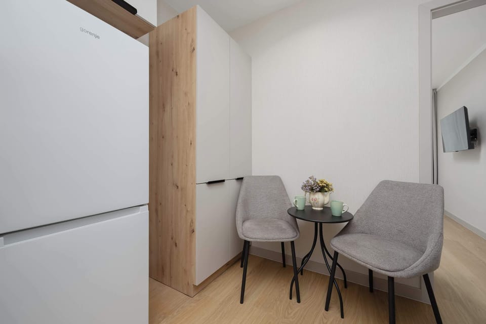 Two grey chairs and a small table are tucked into a corner near a large white refrigerator. This setup provides a functional breakfast spot within a limited floor plan.