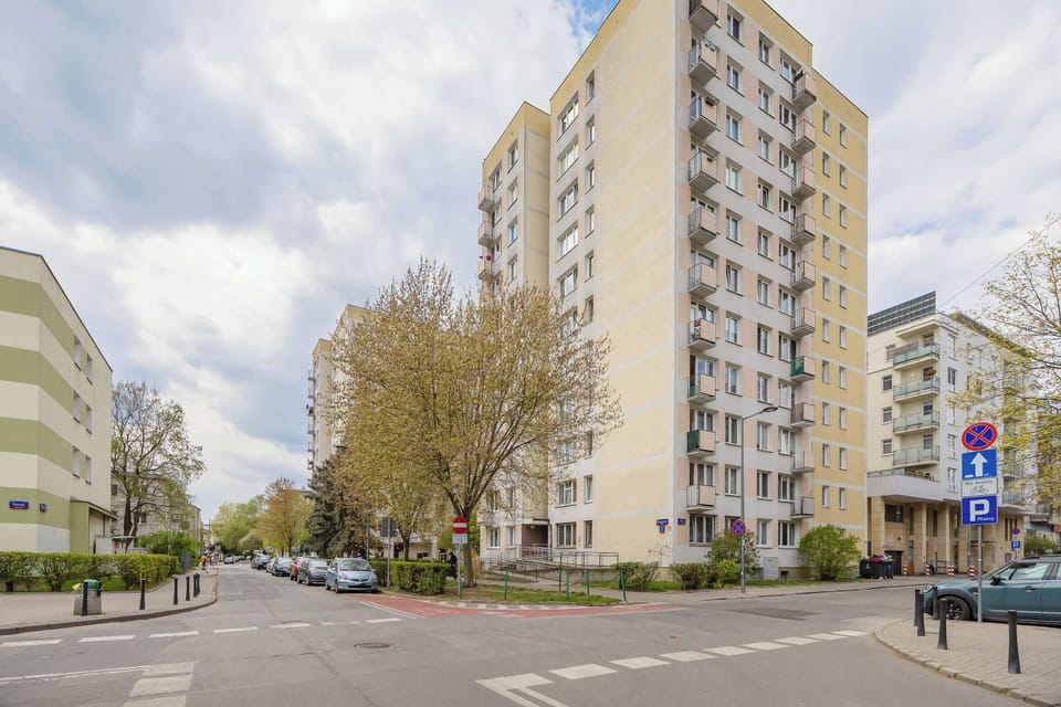 The exterior shot shows a tall, multi-story residential building under a cloudy sky. The building is surrounded by green trees and a paved street, typical of a modern urban neighborhood.