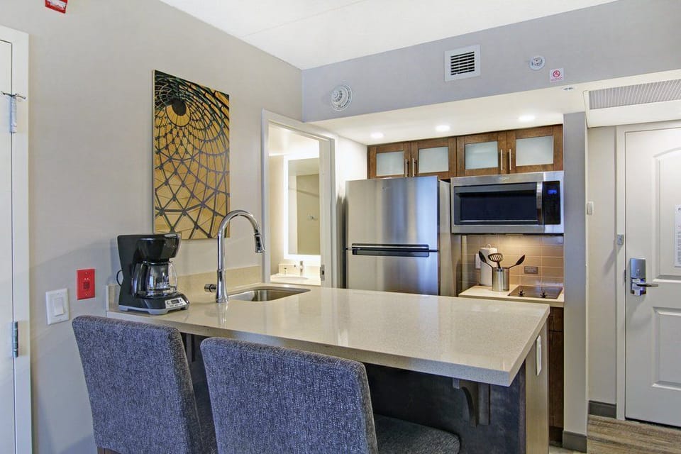 Kitchen bar with two grey upholstered stools, wood cabinets, stainless steel fridge, microwave, and a coffee maker on the counter.