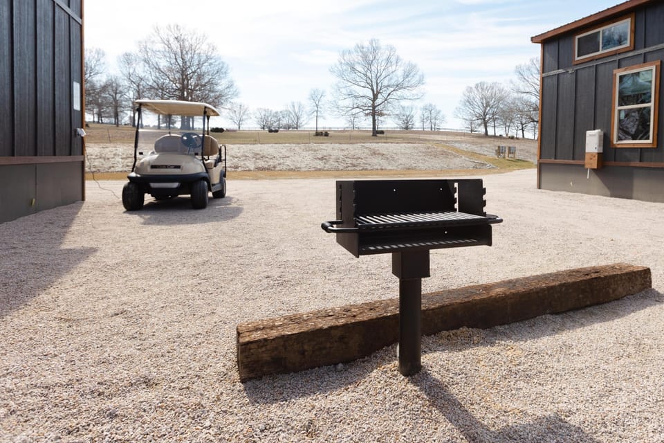 Black metal grill stands on gravel near rustic wooden beam, with golf cart and dark gray cottage exterior in background.