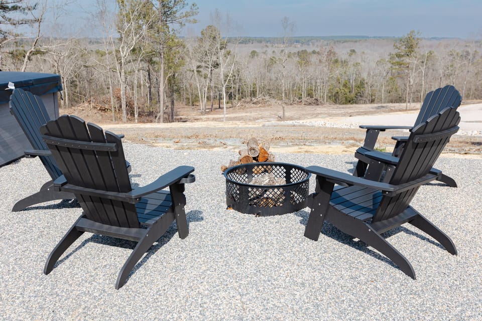 Four black Adirondack chairs circle a metal fire pit on a gravel patio overlooking a wooded landscape.