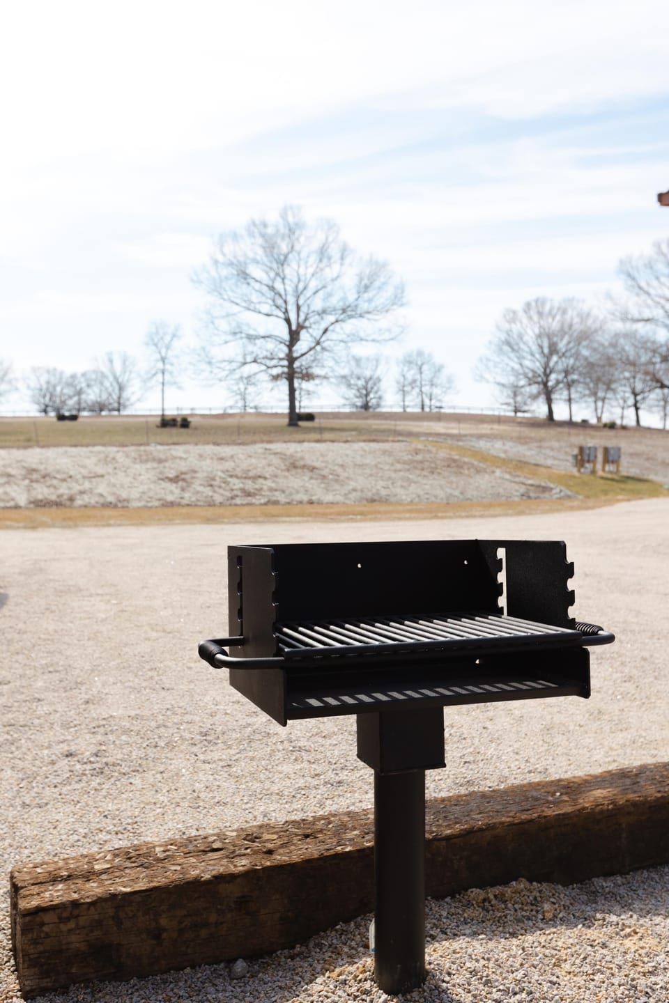 Black metal grill stands on a gravel patio bordered by weathered wooden beams with winter trees in the background.