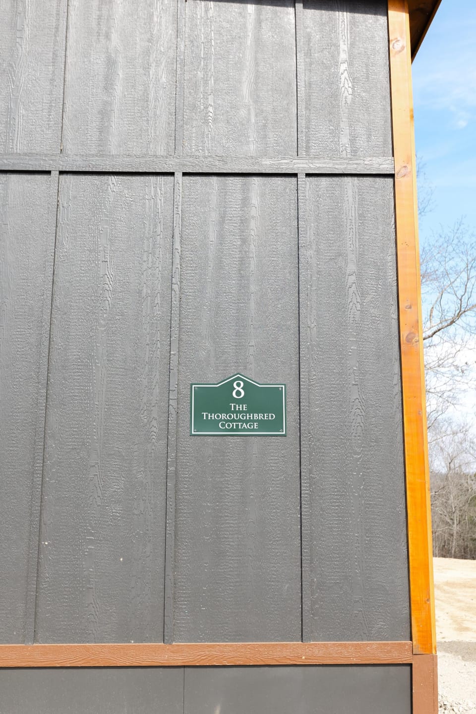 Close-up of dark gray wooden exterior siding with natural wood trim and a green cottage number plaque.