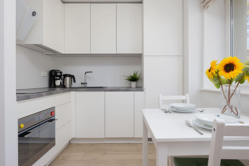 A compact white kitchen features a wooden dining table and two chairs in the foreground. A vase filled with bright yellow sunflowers adds a cheerful pop of color near the window.