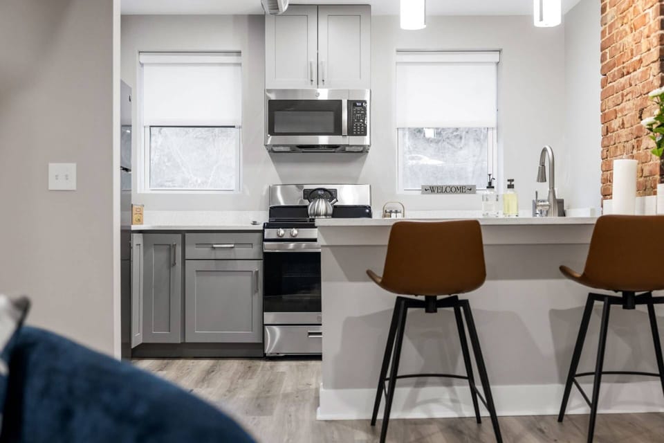 Kitchen from the living area showing stainless appliances, gray shaker cabinets, pendant lighting, and exposed brick.