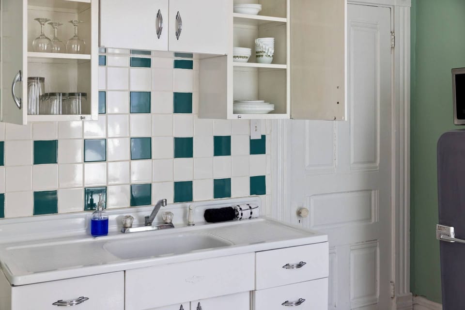 Retro metal sink with original checkerboard tile backsplash and open shelving stocked with glasses and dishware.