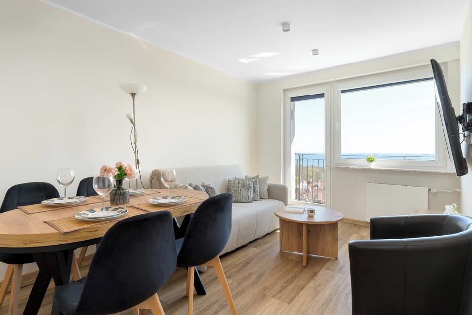 A wider, head-on perspective of the set dining table shows all four modern black chairs arranged neatly. The large patio doors open onto the balcony, offering a direct view of the green landscape and distant water.